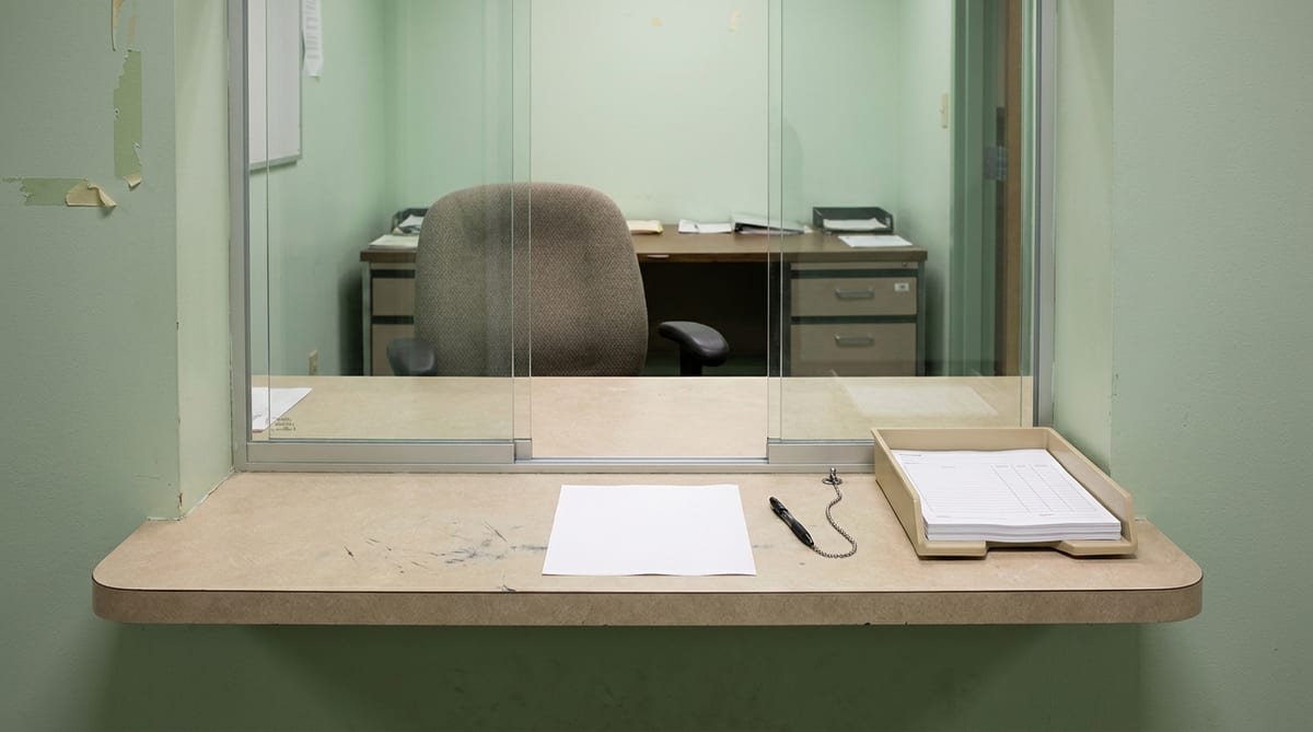 Formulario en blanco sobre un mostrador de trámites, con un bolígrafo encadenado y una silla vacía al otro lado
